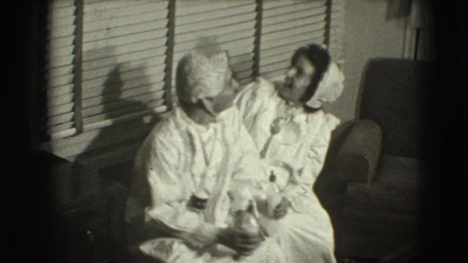The final frame shifts to a domestic interior, where two women in light nightwear sit close together beside window blinds.