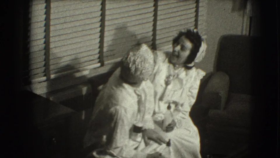 In a Joliet home interior from the 1941 reel, a woman in a white dress and hat sits beside a smaller bonneted figure by closed blinds.