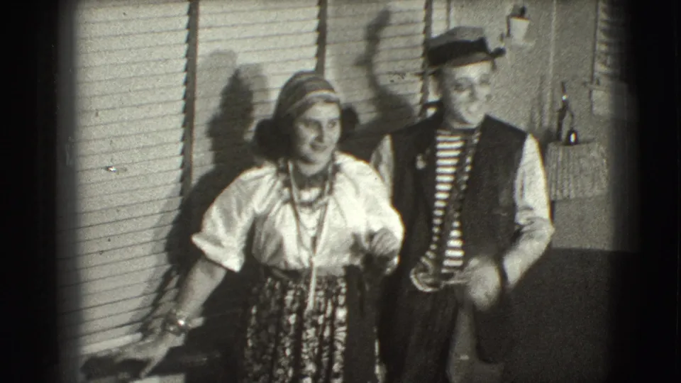 A couple in period-style attire stands together in a Joliet interior, with the woman in a blouse and patterned skirt and the man in a striped shirt and hat.