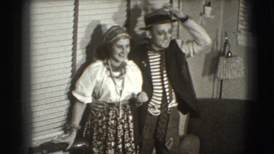 The standing couple smiles for the home-movie camera in the Joliet interior as the man raises a hand to his brimmed hat.