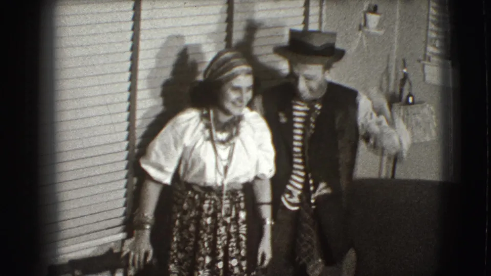 The couple remains close together in the 1941 Joliet home-movie frame, smiling in costume-like clothing near blinds and a wall lamp.