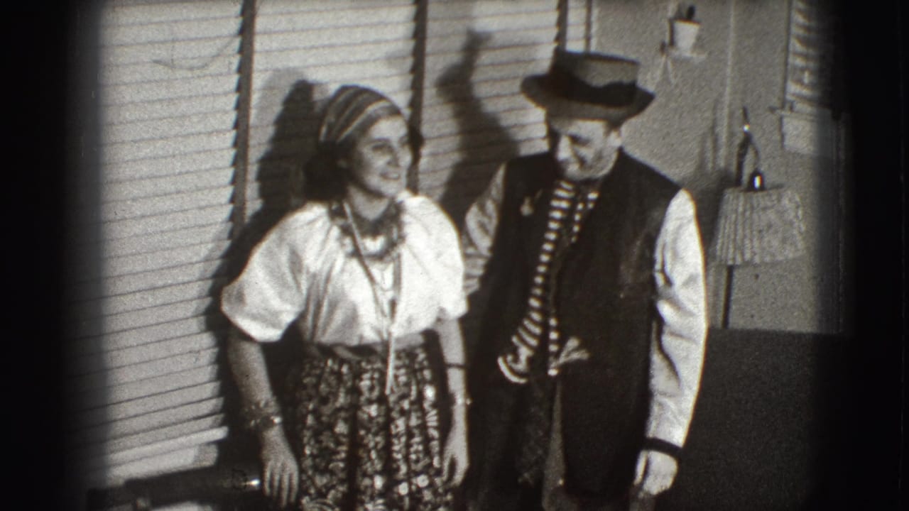 Two costumed adults stand beside window blinds in a home interior.