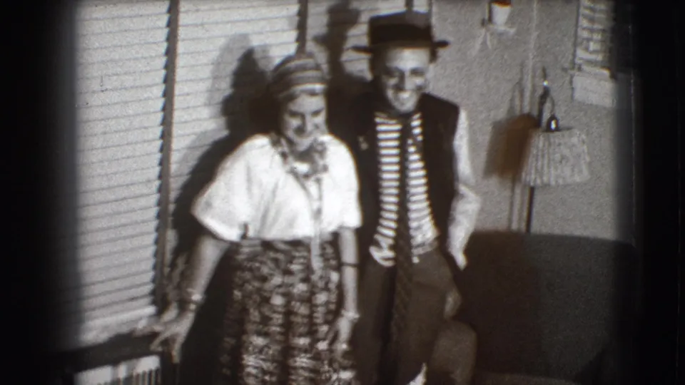 The woman in a headscarf and patterned skirt leans near the window while the man in a fedora smiles behind her in the home interior.