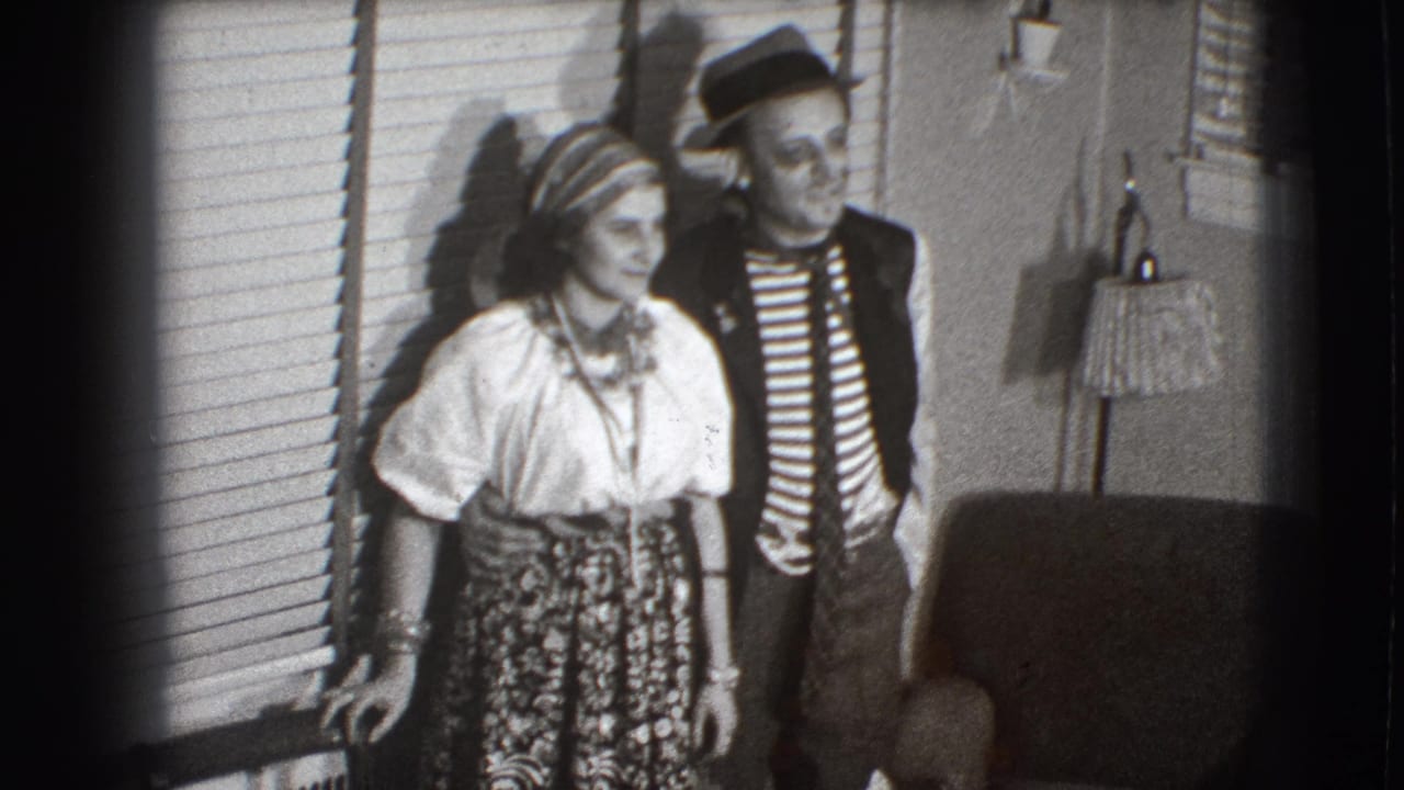 Two adults in costume stand indoors near window blinds and a lamp.