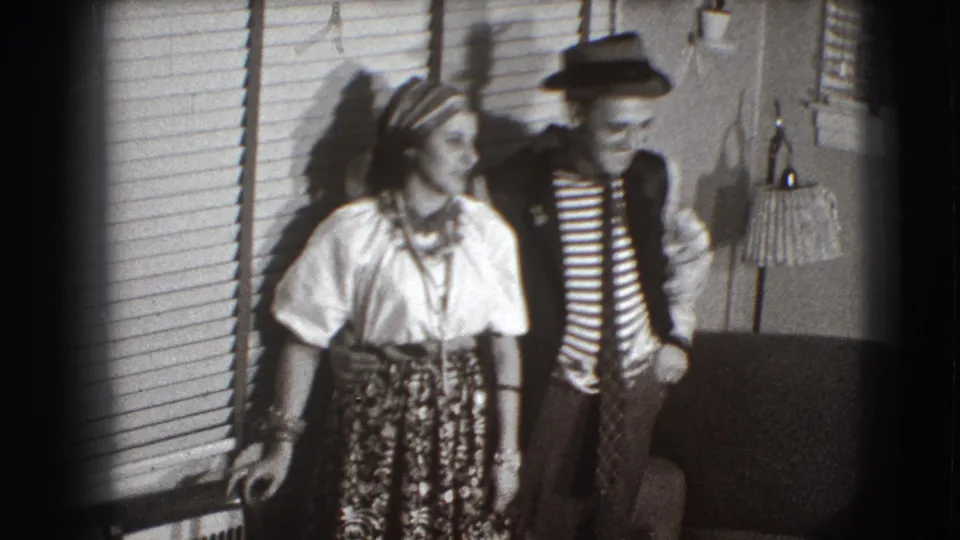 The costumed pair remains posed indoors in the Joliet home-movie frame, with strong shadows falling across the wall and blinds.