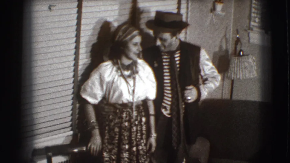 A woman in a blouse and patterned skirt looks toward a man in striped costume clothing inside the Joliet home interior.