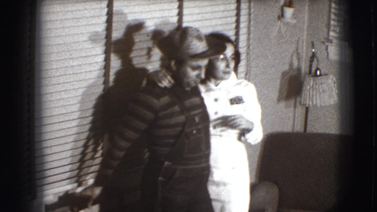 Man in a straw hat stands beside a woman in white in a room with blinds and a lamp.