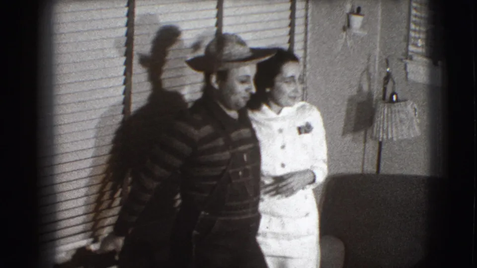 An adult man in a hat and striped shirt stands close to a woman in white inside the Joliet home, framed by blinds and strong shadows.