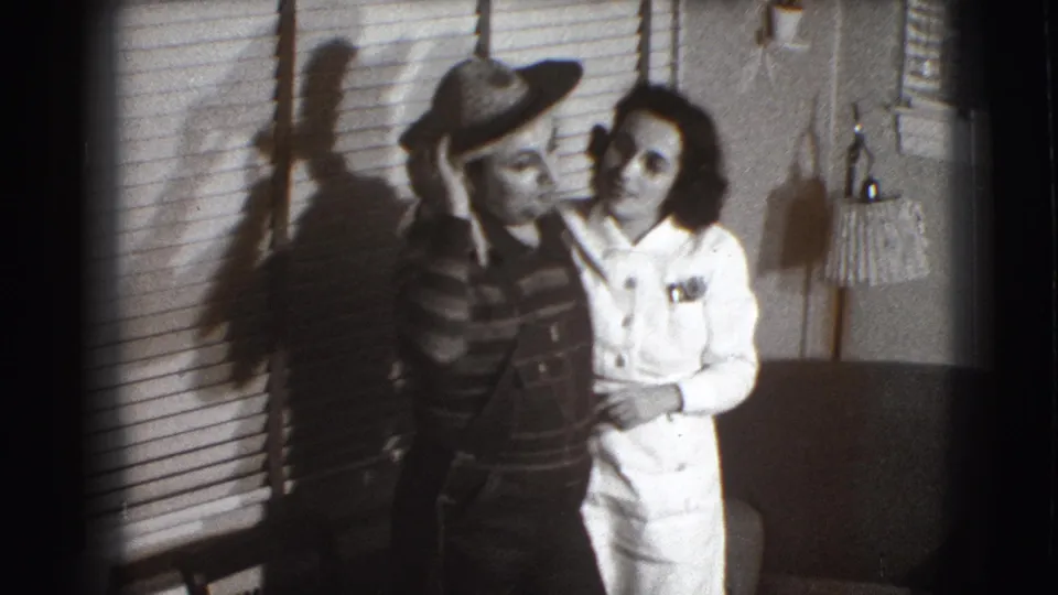 Inside the 1941 Joliet home, the woman leans close while the man raises a hand to his straw hat, with blinds and a chair behind them.