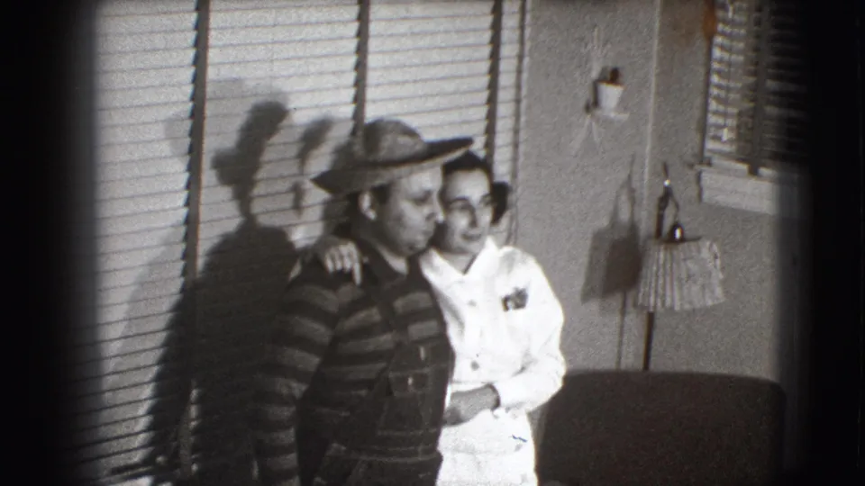 The couple remains close together in the Joliet interior, smiling toward the camera with blinds and a fringed lamp behind them.