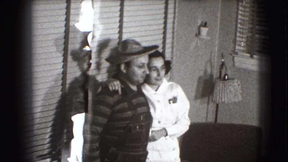 A man wearing a wide-brimmed hat and striped shirt stands beside a woman in a white dress in the 1941 Joliet home interior.