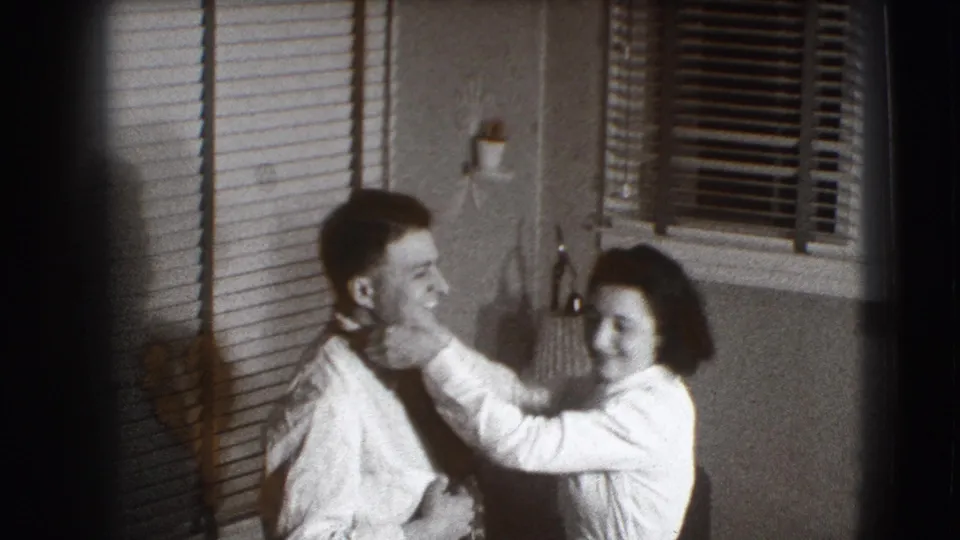 The woman in white reaches toward the man's face as they smile in a private Joliet room with blinds, lamp, and wall sconce visible.