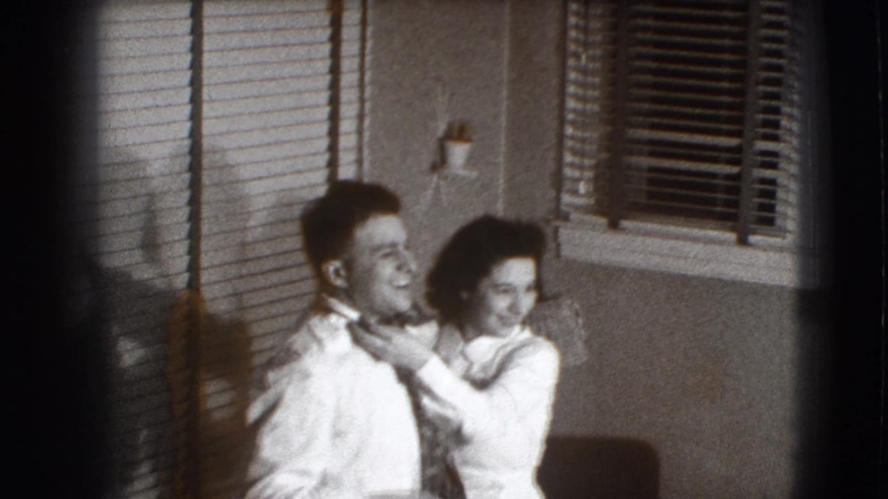 Woman stands behind a smiling man and adjusts his tie beside venetian blinds.