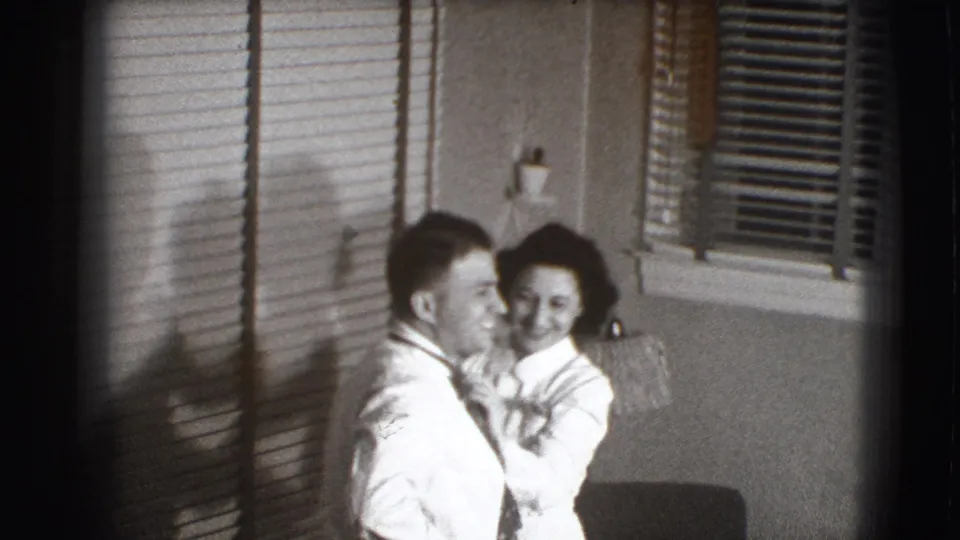 A smiling woman adjusts the man's tie as both stand near horizontal blinds in the 1941 Joliet home-movie scene.
