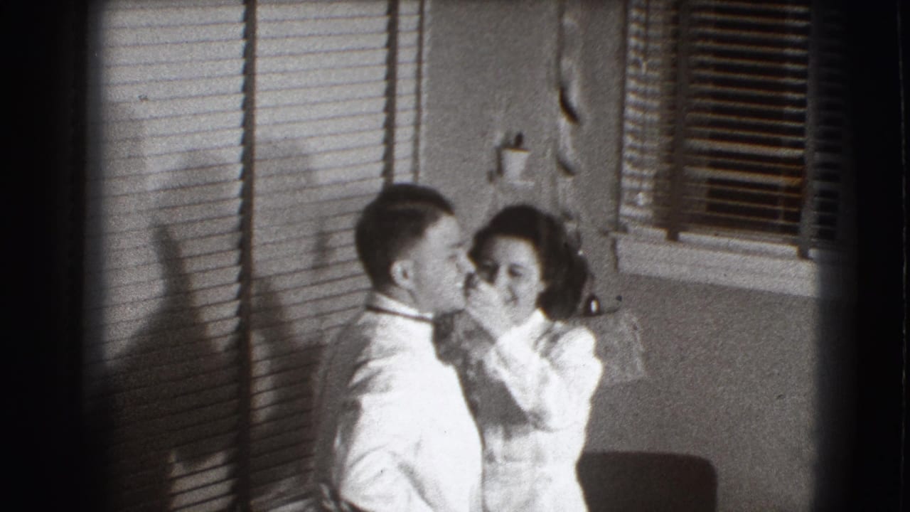 Woman adjusts a man's tie beside window blinds in a home interior.