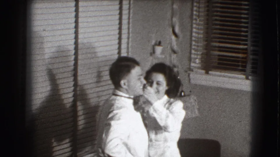 In a 1941 Joliet, Illinois home-movie frame, a woman reaches toward a man's tie as blinds cast striped shadows on the wall.