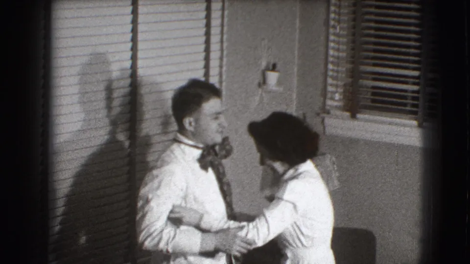 The couple stands in a private Joliet interior as the woman holds the man's arm and works near his tie or shirtfront.