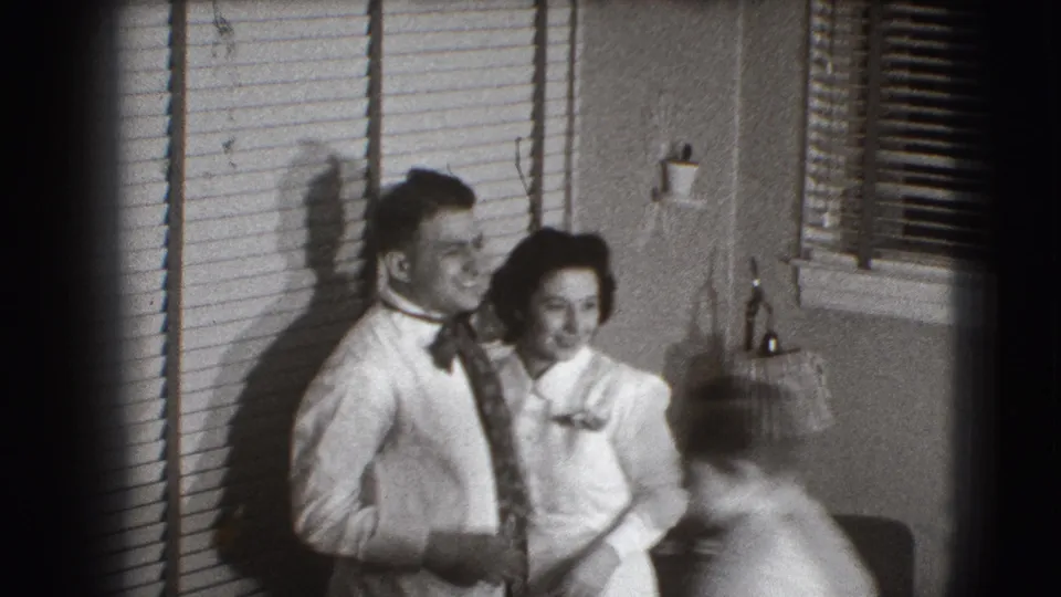 A smiling couple poses near the blinds in the Joliet home interior while another adult figure passes blurred in the foreground.