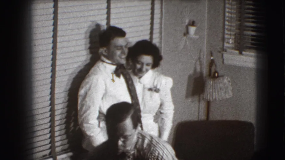In the final frame, the couple remains near the blinds while a seated man appears in the foreground of the private home interior.