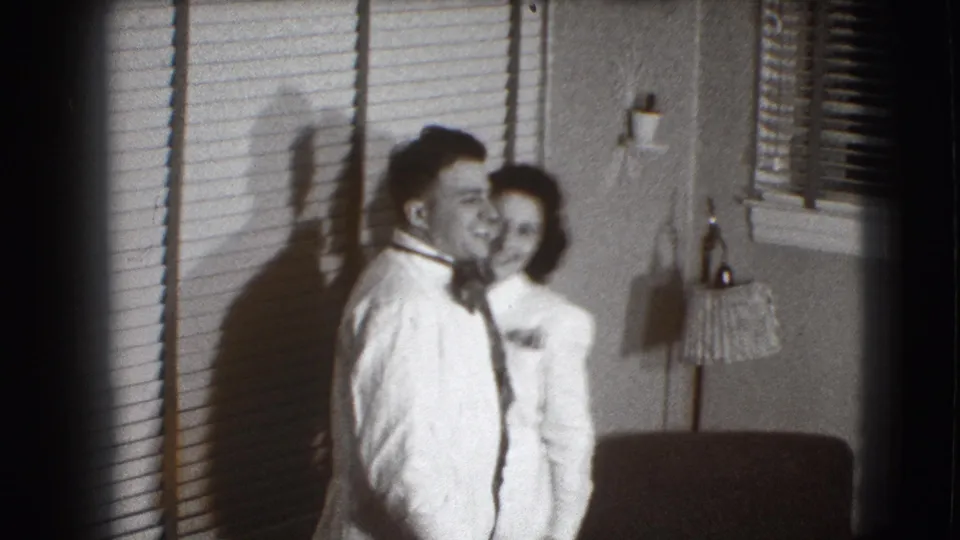 In a 1941 Joliet, Illinois home movie, a young couple smile together indoors beside horizontal blinds and strong wall shadows.
