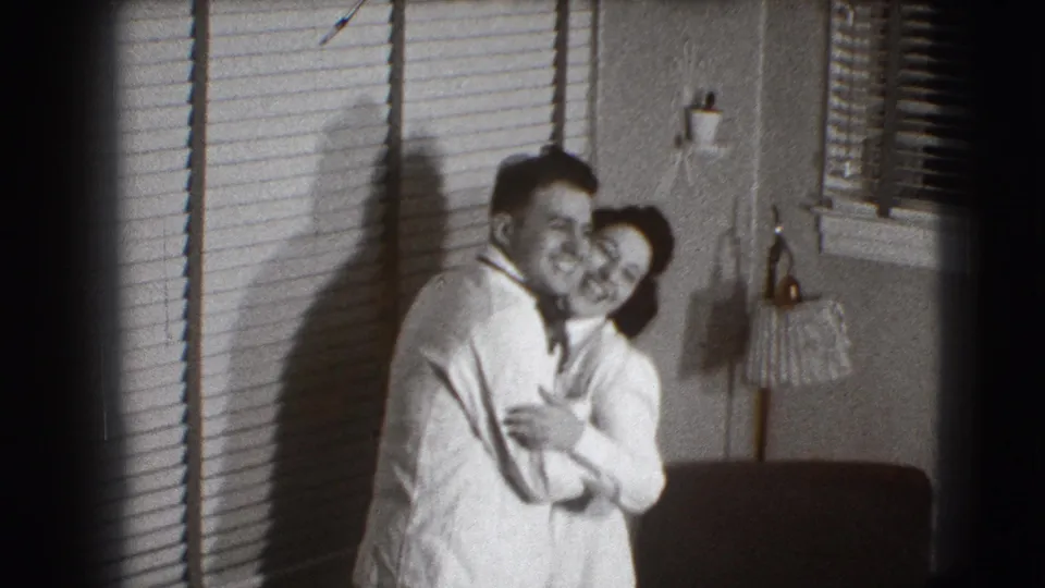 The couple laugh and embrace indoors in the supplied 1941 Joliet home-movie footage, with blinds and a wall fixture behind them.