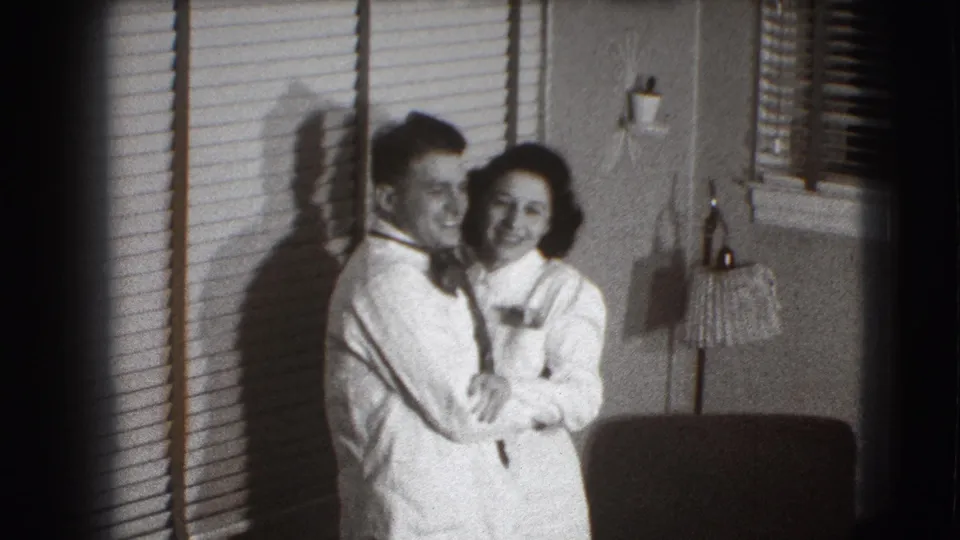 A young couple in formal attire smile close to the camera in a Joliet, Illinois interior, with blinds and shadows behind them.