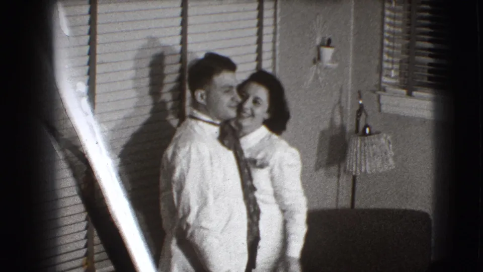 The couple remain side by side in the 1941 home-movie frame, smiling in a domestic room marked by blinds, shadows, and film edge flare.