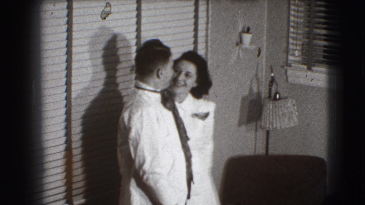 Man in white shirt and tie kisses a woman beside window blinds in a living room.