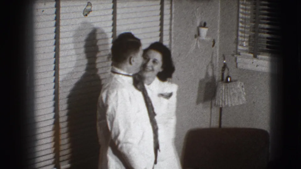 In a 1941 Joliet, Illinois home interior, a man in a white shirt and tie kisses a woman near venetian blinds and a lamp.