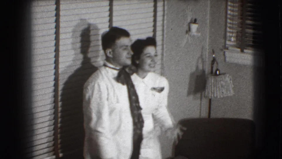 The couple smiles in a Joliet living room, standing close by venetian blinds with a shaded lamp and furniture visible.
