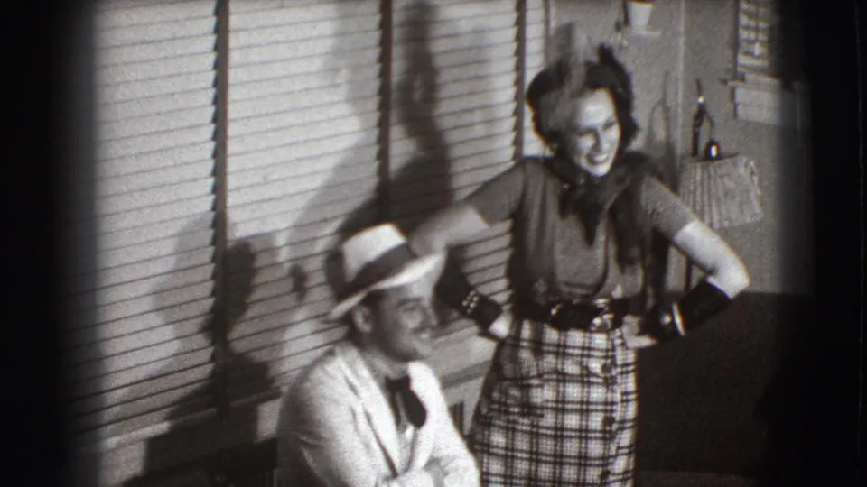 The woman poses above a seated man in a light hat, framed by venetian blinds, a lamp, and living-room furniture in Joliet.