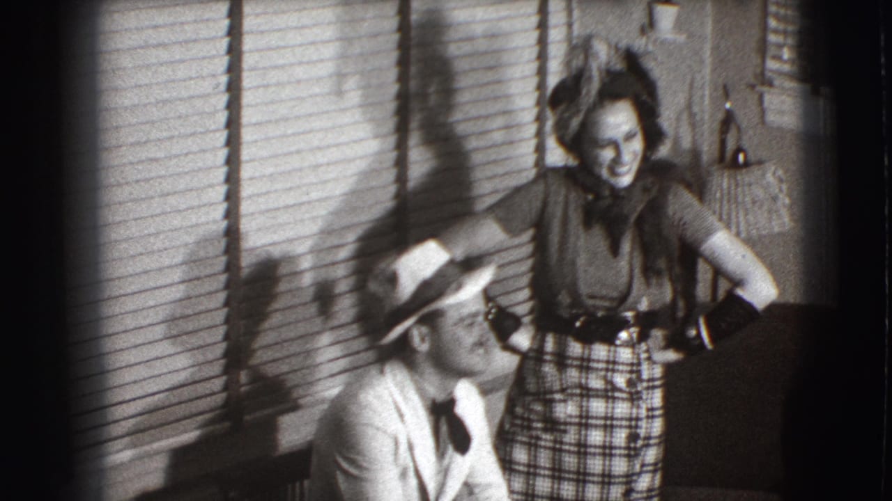Seated man in a light suit and fedora smiles beside a laughing woman in a plaid skirt indoors.