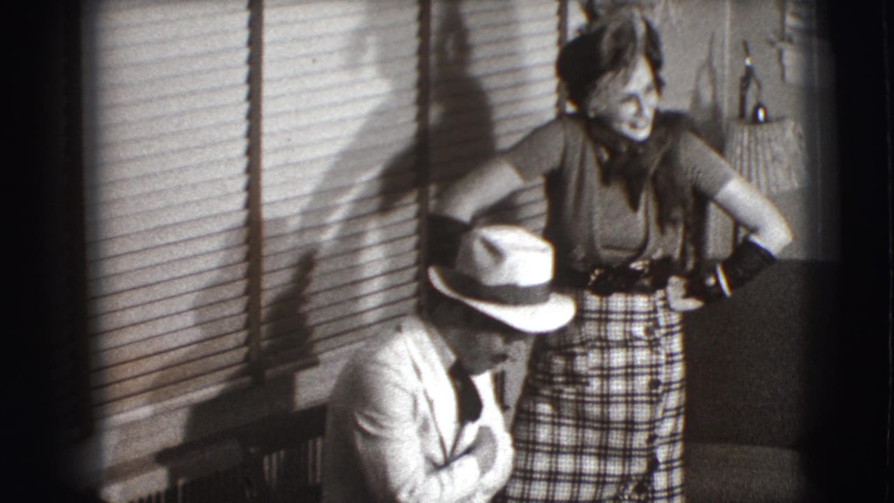 Seated man in a fedora beside a standing woman in a plaid skirt indoors.