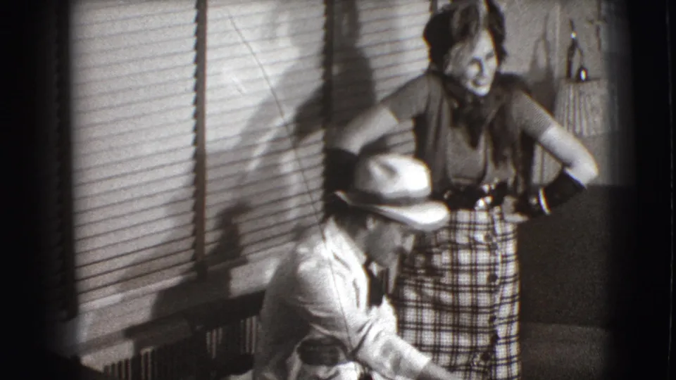 The Joliet home-movie frame shows the woman smiling over a seated man in a fedora beside blinds and interior furnishings.