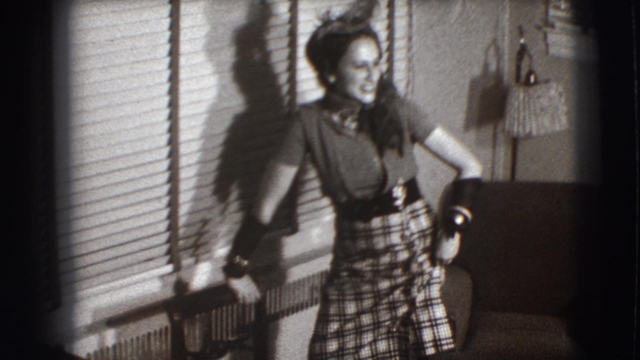 Young woman in a plaid skirt laughs while leaning near window blinds in a home interior.
