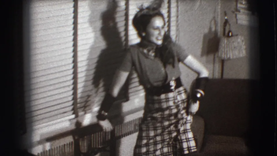 In 1941 Joliet home-movie footage, a young woman in a plaid skirt and hat laughs beside window blinds and an armchair.