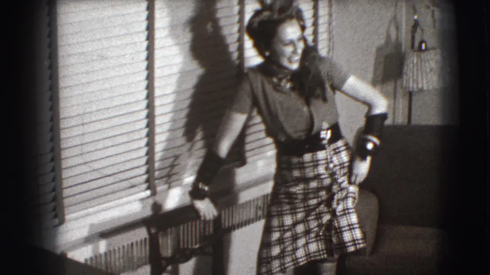 The woman continues posing in the Joliet interior, smiling beside venetian blinds, a radiator, an upholstered chair, and a lamp.