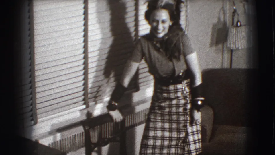 A young woman smiles toward the camera in the 1941 Joliet interior, with blinds, radiator, armchair, and lamp behind her.