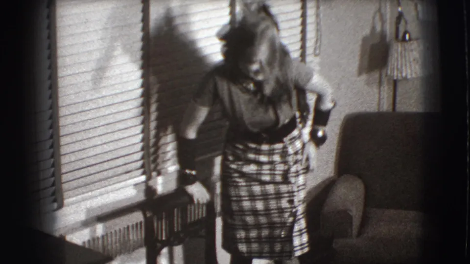 At the end of the clip, the woman bends forward laughing in the Joliet room, with window blinds, radiator, chair, and lamp visible.