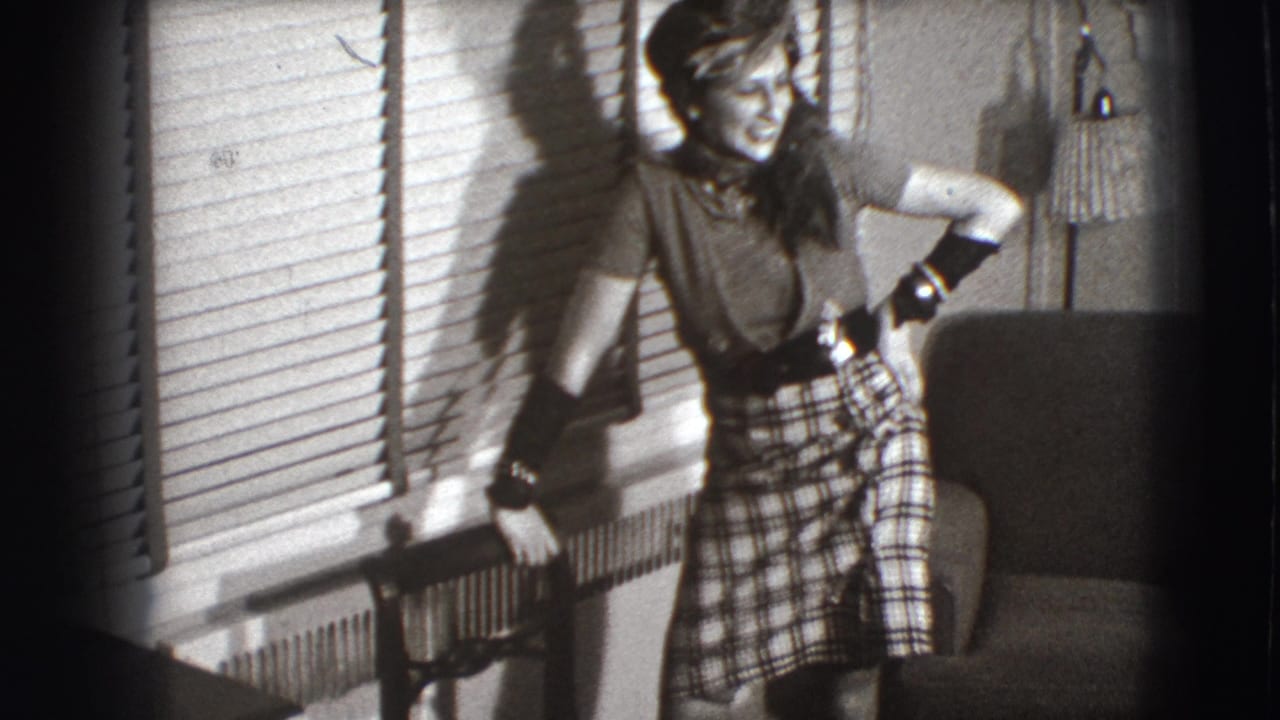 Woman in a feathered hat and plaid skirt stands beside venetian blinds in a living room.