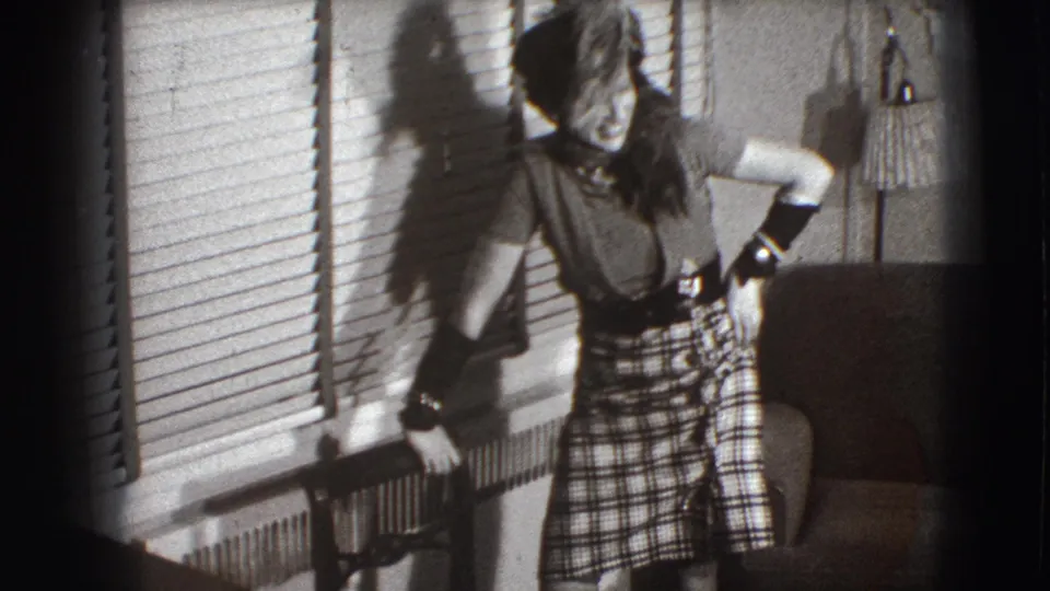 The woman remains near the venetian blinds in the Joliet interior, with her plaid skirt, gloves, radiator, chair, and lamp visible.