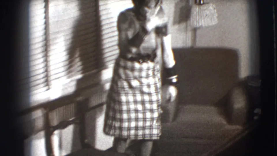 In 1941 Joliet home-movie footage, a woman in a plaid skirt stands in a sunlit living room near venetian blinds and an armchair.