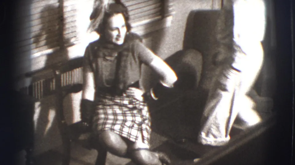 The woman sits in the living room near an armchair and window, with another adult figure partly visible at the edge of the frame.