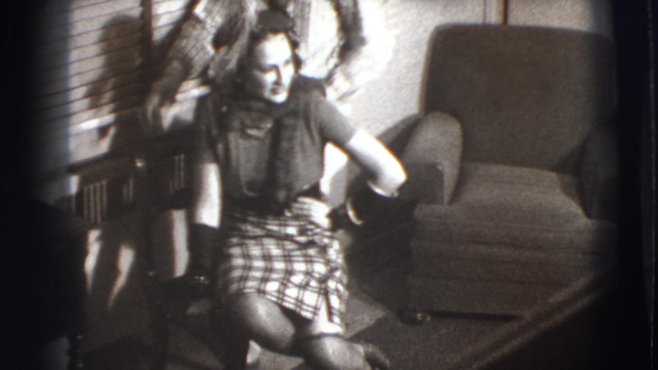 Seated woman in plaid skirt smiles in a living room as another person stands partly behind her.