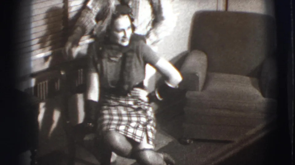 In a 1941 Joliet home-movie frame, a woman sits smiling in a living room with blinds and an armchair while a person stands partly behind her.