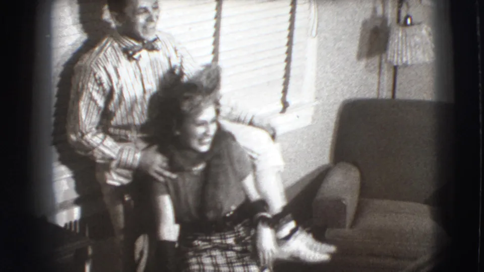 In a 1941 Joliet living-room frame, a man in a striped shirt and bow tie stands behind a smiling seated woman near blinds and an armchair.