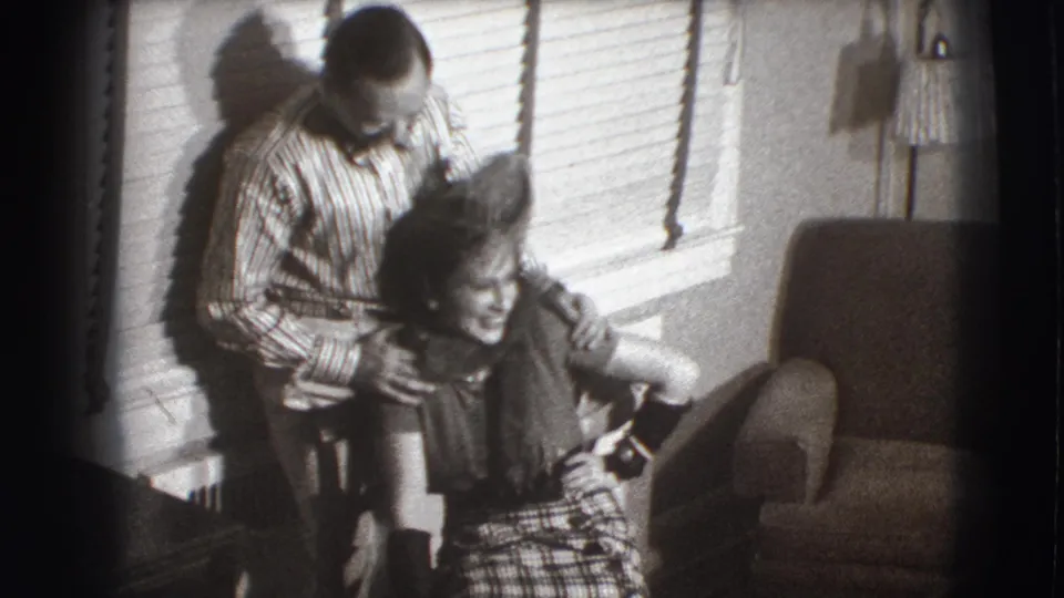 A 1941 Joliet home-movie frame shows a man leaning over a seated woman, his hands near her shoulders, as she smiles in a living room.