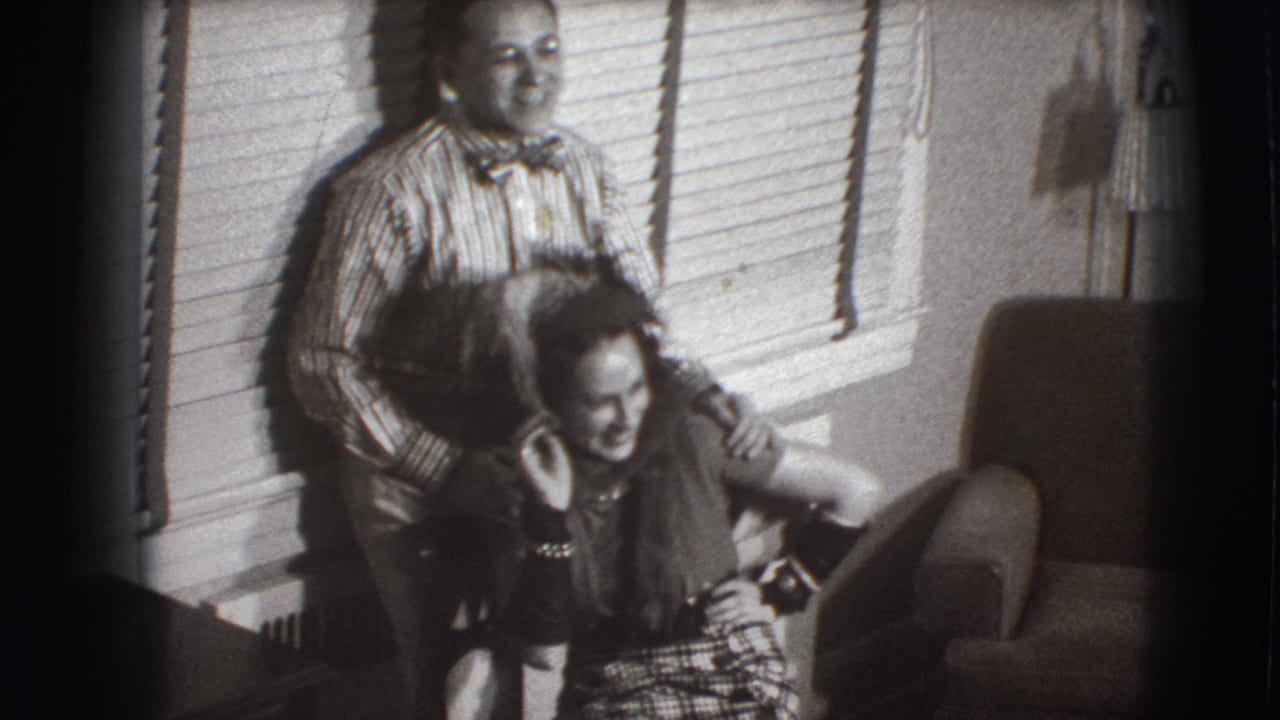 Man in striped shirt stands behind a seated woman laughing in a living room.