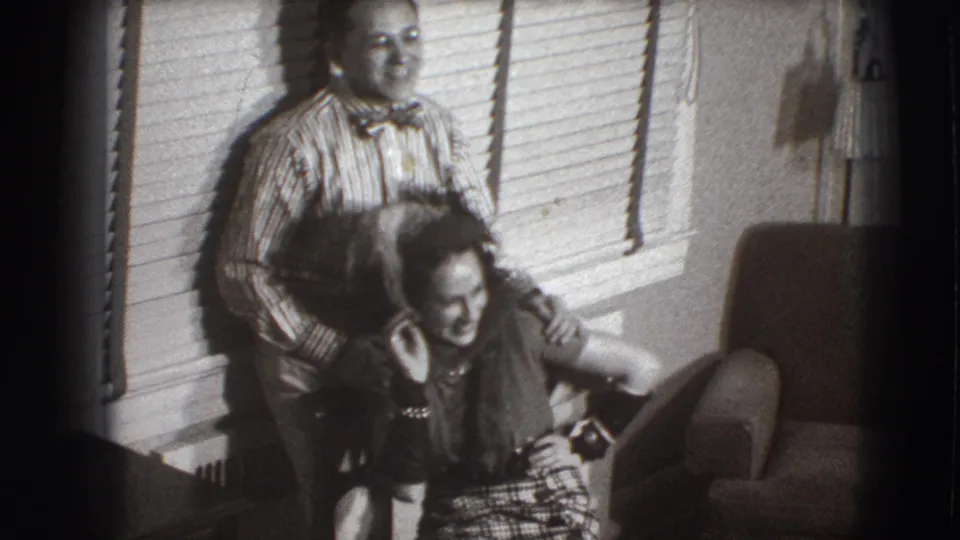 In a Joliet, Illinois 1941 home-movie frame, a man in a striped shirt and bow tie stands behind a seated laughing woman.