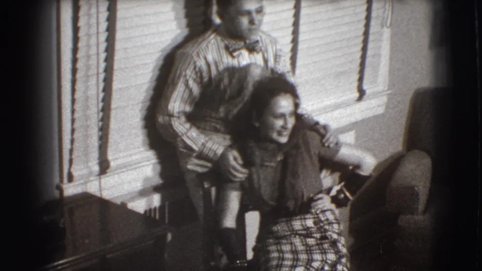 A black-and-white 8mm frame shows the seated woman laughing in a plaid skirt as the man stands close behind her in the living room.